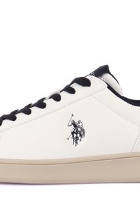 Baskets blanches avec lacets noirs et semelle beige, avec un logo joueur de polo noir sur le côté.