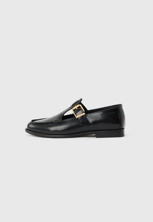 Black leather loafer with gold buckle strap, moc toe stitching, and low flat heel, side view on neutral background.
