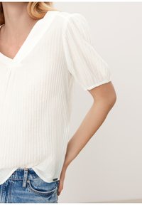 White, textured blouse with a V-neck, puffed short sleeves, and a hemline that drapes slightly over blue denim jeans.