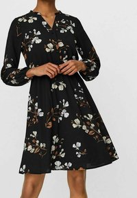 Musta kukallinen mekko pitkillä hihoilla, jossa on nappisulku, valkoisia ja ruskeita kukkakuvioita sekä virtaava A-linjainen siluetti.
