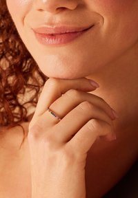 Gouden ring met kleine, kleurrijke edelstenen gezet in een delicate band. Een hand rust nabij de kin, waardoor het ontwerp en de glanzende afwerking van de ring benadrukt worden.