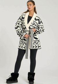 Knittröja med svartvita geometriska mönster, sjalkrag, bälte i midjan och långa ärmar, i kombination med svarta leggings och fluffiga stövlar.