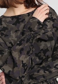 Camo-mönstrad topp i mörkgrön och grå nyanser. Har breda ärmar med samlad detaljer. Mjuk, texturerad tyg. Rund halsringning.