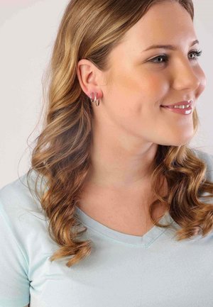 Boucles d'oreilles en argent en forme de créoles avec trois anneaux empilés ; le modèle a des cheveux ondulés jusqu'aux épaules et porte un t-shirt bleu clair à col en V.