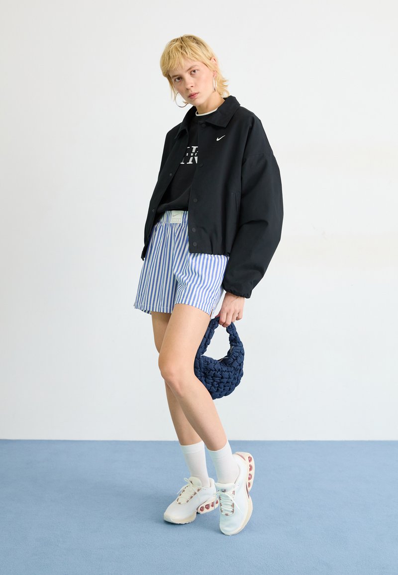 Veste bomber noire, shorts rayés, baskets blanches avec accents colorés, et un sac à main texturé marine. Pose à plat sur fond bleu clair.