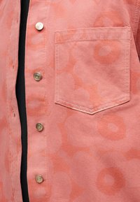 Κοντινό πλάνο πουκάμισου με ροζ λουλουδάτο σχέδιο, μεταλλικά κουμπώματα και τσέπη στο στήθος πάνω από μαύρο ρούχο.