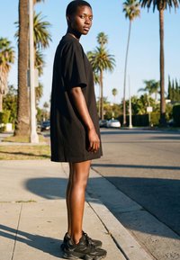 Fiatal nő fekete, túlméretezett pólóban és sportcipőben, napfényes járdán áll, háttérben pálmafák és autók.