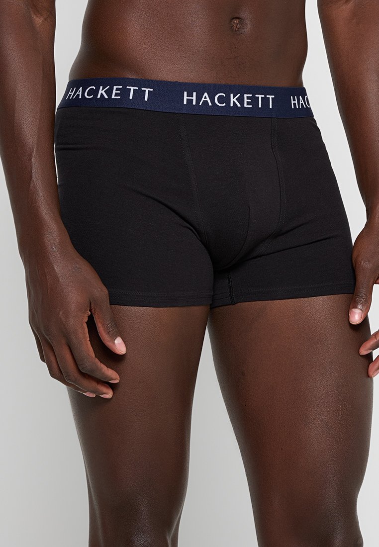 Boxer ajustados de algodón negro con una cinturilla elástica en azul marino que presenta el texto blanco "HACKETT". Textura suave, diseño ajustado y longitud a mitad del muslo.