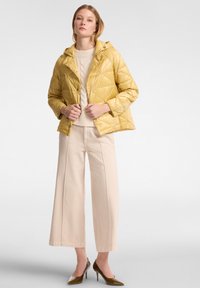 Giacca trapuntata gialla con cappuccio, realizzata in un materiale resistente e con chiusura frontale, abbinata a un maglione a maglia beige e pantaloni larghi color beige chiaro.