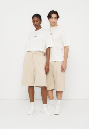 Twee modellen staan zij aan zij in witte oversized T-shirts, beige lange shorts, witte sokken en witte sneakers tegen een effen witte achtergrond.