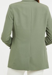 Blazer vert olive avec une texture lisse, coupe ajustée et fente arrière. Présente des revers crantés et des manches longues, conçu pour une silhouette cintrée.
