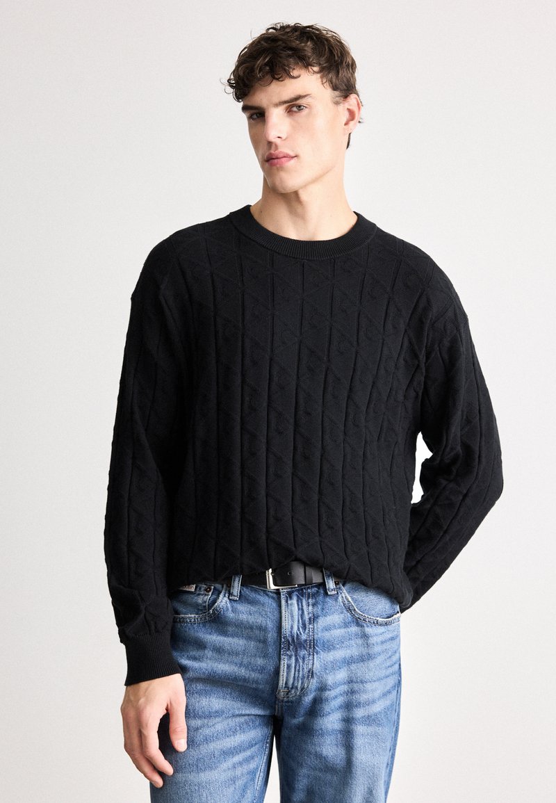 Maglione nero lavorato a maglia con motivo a diamante, polsini e scollatura a costine, abbinato a jeans azzurri chiari. La vestibilità è comoda.