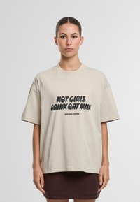 Camiseta de algodón beige con corte oversize, mangas cortas, que presenta un texto negro en negrita "NO CHICAS BEBEN LECHE DE AVENA" y el nombre de la marca debajo.