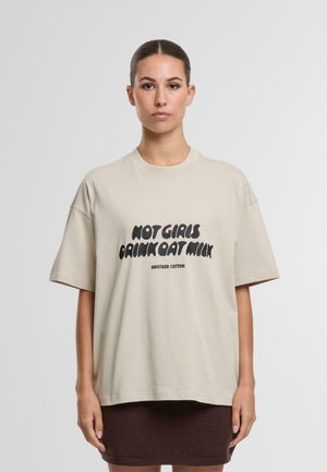 Camiseta de algodón beige con corte oversize, mangas cortas, que presenta un texto negro en negrita "NO CHICAS BEBEN LECHE DE AVENA" y el nombre de la marca debajo.