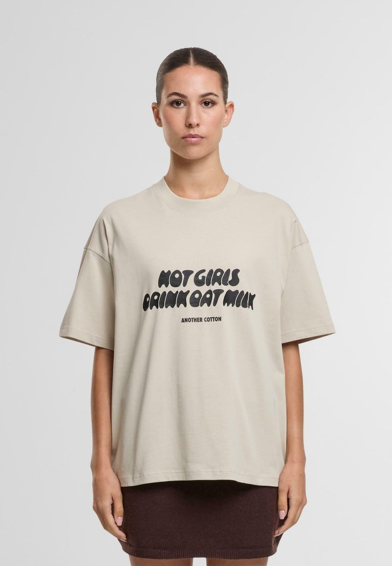 Camiseta de algodón beige con corte oversize, mangas cortas, que presenta un texto negro en negrita "NO CHICAS BEBEN LECHE DE AVENA" y el nombre de la marca debajo.