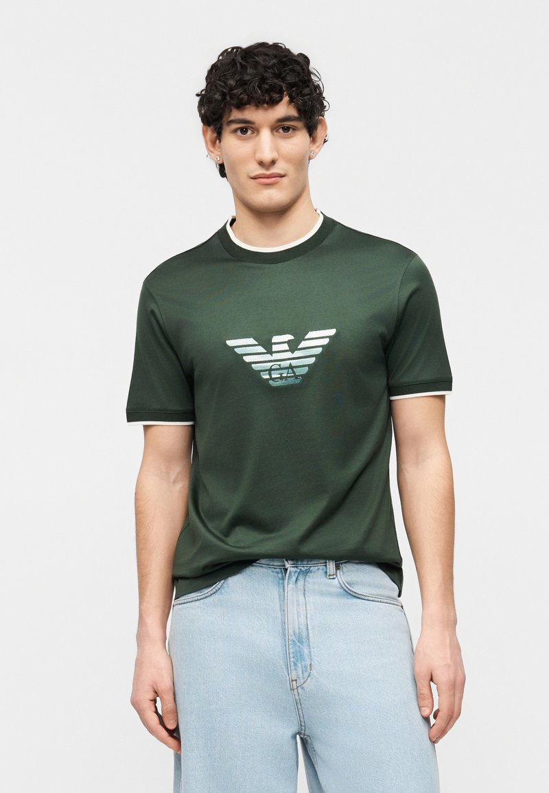 Νεαρός άνδρας με σγουρά μαλλιά, φορώντας κοντομάνικο πουκάμισο σκούρου πράσινου χρώματος με λευκή διακόσμηση και λογότυπο, συνδυασμένο με ανοιχτόχρωμο τζιν παντελόνι.