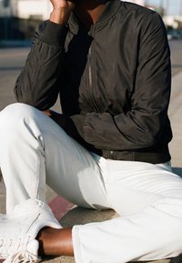 Personne portant une veste noire zippée, un pantalon blanc et des baskets blanches, assise à l'extérieur avec un genou levé et la main reposant près du visage.