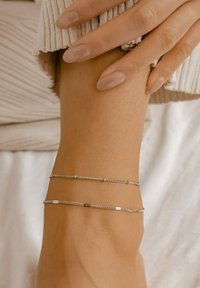 Pulsera de doble cadena ajustable de plata con eslabones cilíndricos, que realza el diseño elegante. Mano apoyada sobre una superficie texturizada.