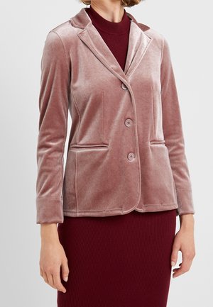 Vrouw draagt een stofroze velvet blazer met knopen over een bordeauxroze geribbelde jurk, staand tegen een effen lichte achtergrond.