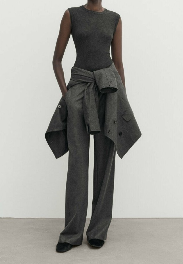 Personne portant un haut sans manches gris foncé et un pantalon large assorti, avec une veste grise attachée autour de la taille, debout devant un fond uni.