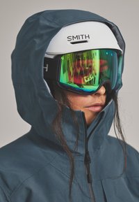 Ski helmet with a white shell, branded "SMITH," paired with green-tinted ski goggles. Hooded blue jacket with a zip closure and textured finish.
