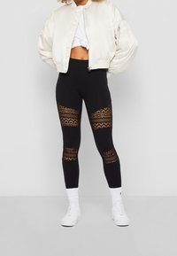 Leggings noirs avec des panneaux en dentelle transparente et un design ajusté, associés à une veste bomber crème. Des baskets blanches complètent la tenue.