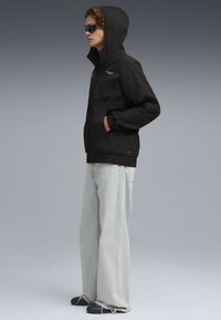 Chaqueta ligera negra con capucha, con un logo verde, combinada con jeans sueltos de mezclilla azul claro y zapatos negros.