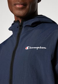 Navy hooded jacket made of lightweight fabric, featuring a front zipper and white "Champion" logo with a red accent.