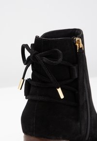 Botín de ante negro que presenta una cremallera lateral y detalles de cordones con acentos dorados. El diseño incluye un material texturizado y un tacón de madera bajo.