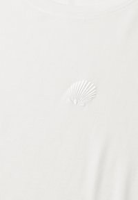 Tissu blanc avec un petit motif de coquillage brodé en fil blanc centré près de la zone supérieure.