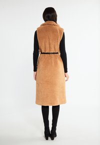 Langer brauner Kunstpelz-Westen mit hohem Kragen, tailliertem Schnitt und ärmellosem Design. Kombiniert mit schwarzen Leggings und Ankle Boots.