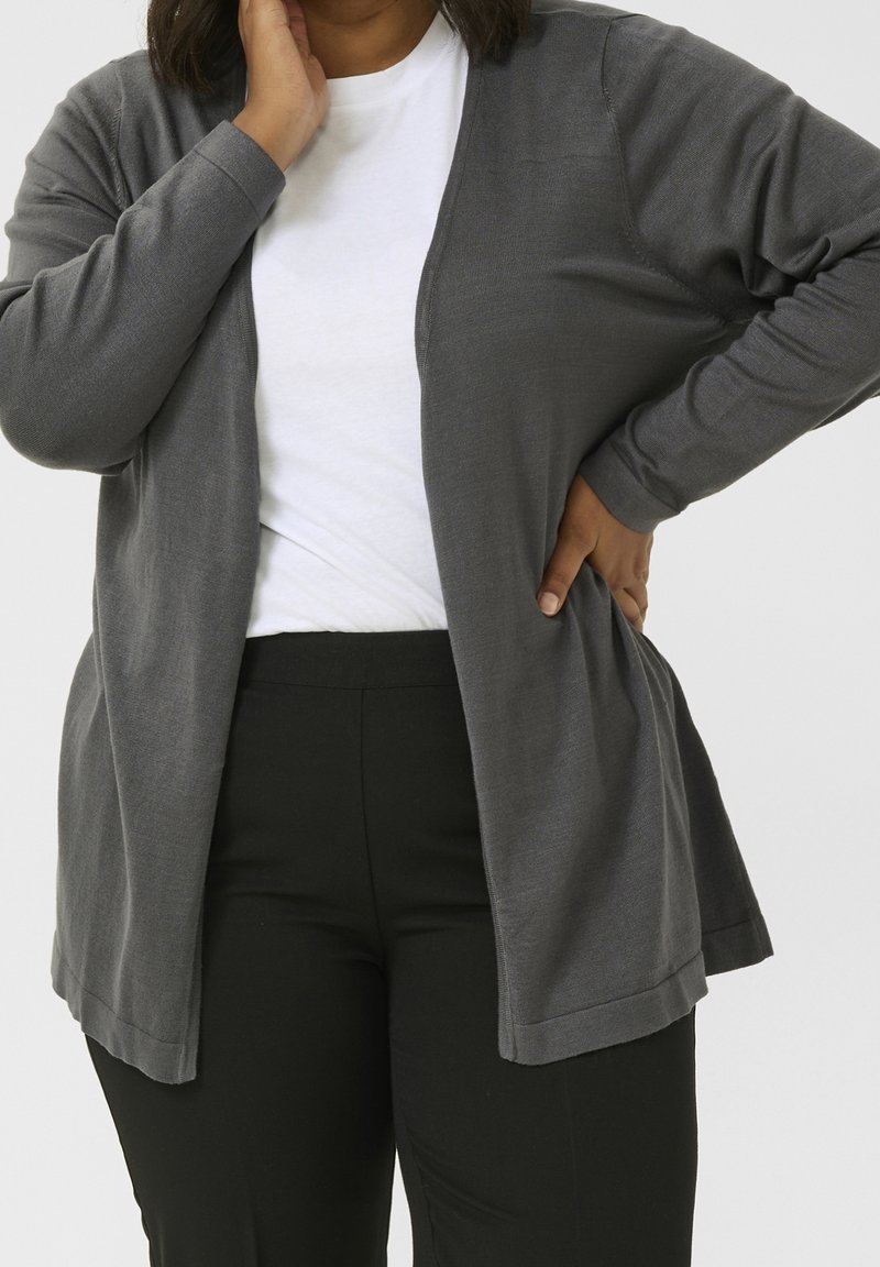 Personne portant une chemise blanche, un cardigan gris foncé ouvert devant et un pantalon noir, une main sur la hanche et l'autre touchant son visage.
