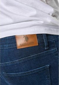 Mörkblå denimjeans med en brun läderlogotyp som är märkt "U.S. Polo Assn." med sömnadsdetaljer och bakfickor synliga.