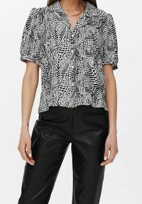 Blouse noire et blanche à manches courtes avec un motif géométrique, un col et une coupe décontractée, associée à un pantalon en cuir noir.