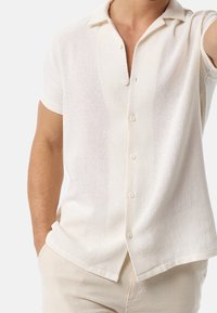 Short-sleeve white woven shirt with a collar, featuring a button front, textured fabric, and straight hem.