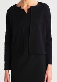 Svart kort jacka med långa ärmar, texturerad tyg med ett subtilt mönster, och rund halsringning, bärs över en svart klänning.