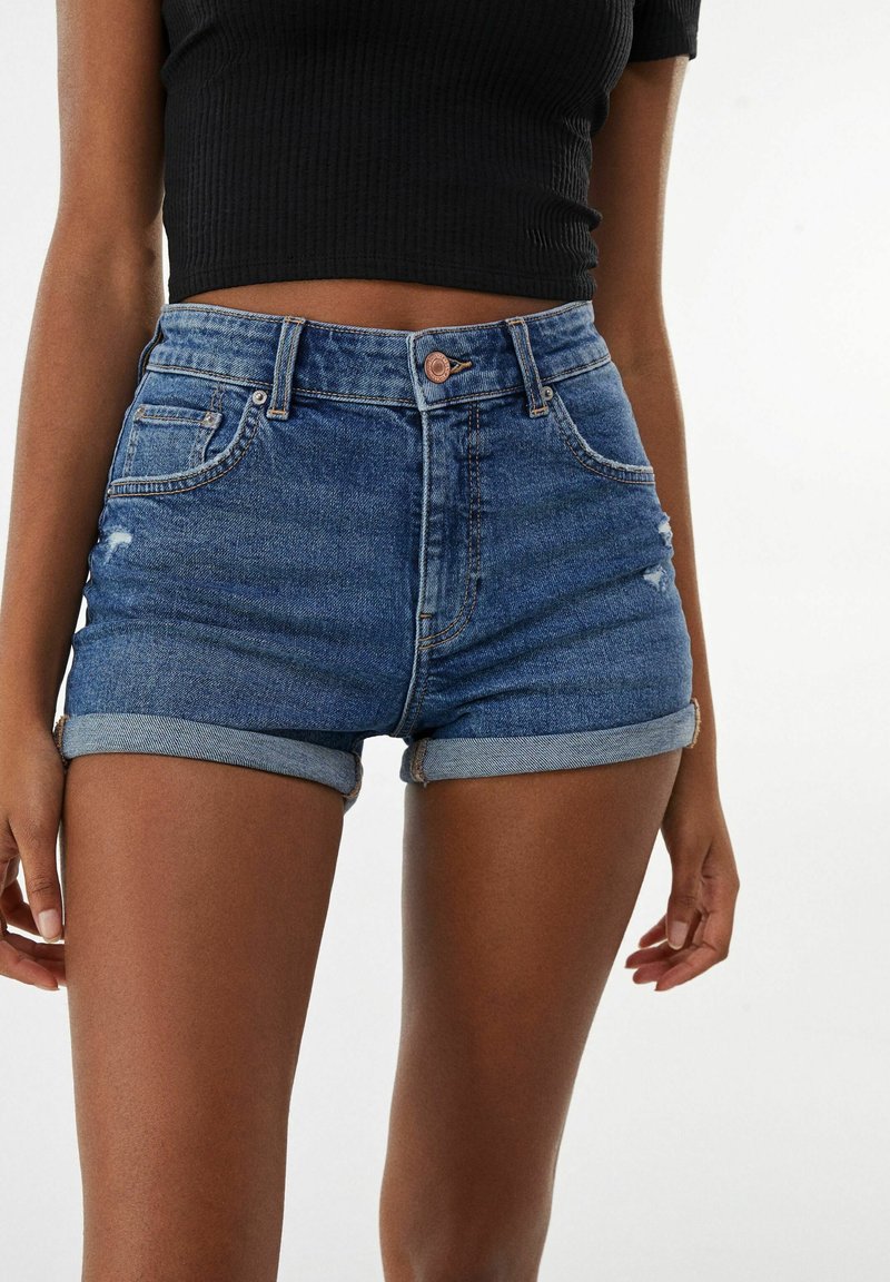 Shorts di jeans in blu medio con orli risvoltati, vita alta, tasche frontali e lievi effetti di usura sulla coscia sinistra.