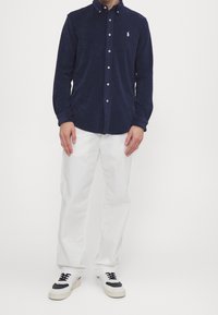 Marinblå långärmad skjorta med knappar, tillverkad av mjukt tyg. Bärs med vita byxor och svart-vita sneakers. Har en liten logotyp på bröstet.