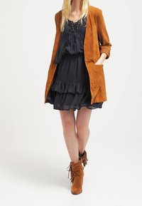 Sammetsbrun jacka, lager på lager över en marinblå klänning med volanger, tillsammans med bruna fransade ankelstövlar. Slät textur, avslappnad passform.
