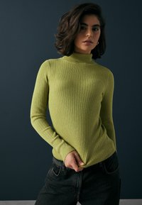 Gerippter, enganliegender Rollkragenpullover in leuchtendem Grün, versehen mit langen Ärmeln und einer strukturierten Oberfläche, kombiniert mit dunklen Jeans.