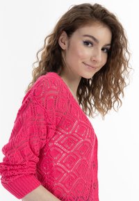 Mönstrad kofta i livlig rosa med intrikata diamantmönster, knäppning med knappar och texturerade ärmar. Lätt och löst vävd design.