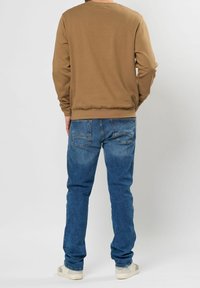Sudadera marrón con puños y dobladillo acanalados, combinada con vaqueros azules que tienen bolsillos traseros y un acabado desgastado, llevada con zapatillas claras.