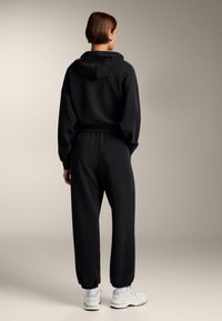 Čierna mikina s voľnými ramenami a elastickými manžetami, kombinovaná s nohavicami na jogging s naťahovacími členkami. Oblečené s bielymi teniskami.