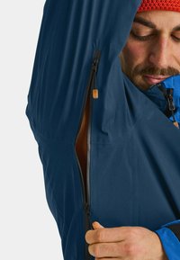 Marineblaue wasserdichte Jacke aus strukturiertem Stoff. Verfügt über einen halben Reißverschluss und kontrastierende blaue Akzente. Sichtbares Innenfutter in Orange.