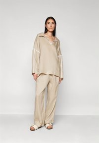 Bež bež majica iz lanenega materiala z V-izrezom in belimi obrobami, združena z ohlapnimi hlačami istega materiala in ravnimi sandali, postavljena na preprostem ozadju.