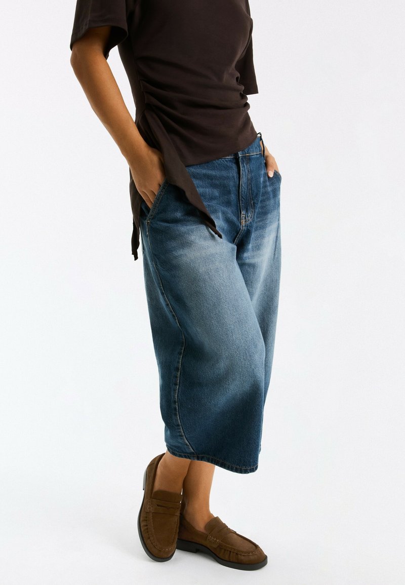 Personne portant une jupe midi en jean bleu, un top noir avec des liens sur les côtés, et des mocassins marron, debout les mains dans les poches sur fond blanc.