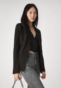 Svart blazer med skarpe klaffer, kontrasterende sømmer, enkel knappelukking og grå skinny jeans. Metallisk kjedeveske holdt i hånden.