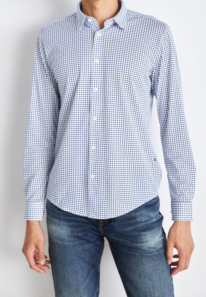Homme portant une chemise blanche boutonnée avec un motif à carreaux bleu et un jean bleu délavé, debout devant un fond blanc uni.