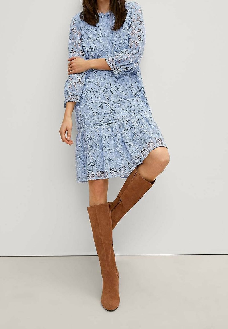 Femme portant une robe en dentelle bleu clair avec des manches trois-quarts et des bottes en daim marron jusqu'aux genoux, posant devant un mur blanc.