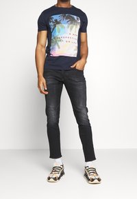 Marinblå t-shirt med en färgglad grafik som föreställer palmer och text; kombinerad med mörka jeans och mönstrade sneakers.